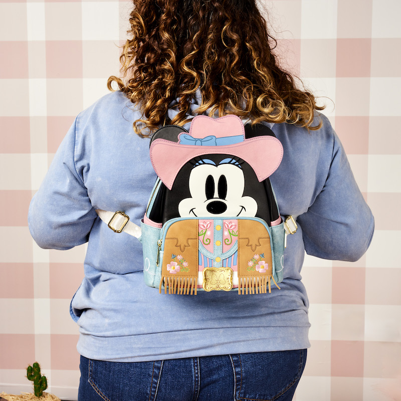 Woman wearing the Western Mickey Mouse Unisex Hoodie facing away from camera and wearing the Loungefly Western Minnie Mouse Cosplay Mini Backpack, against a pastel pink checkered background 